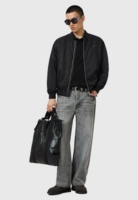 Jeune homme portant une veste bomber noire, une chemise noire, des lunettes de soleil surdimensionnées, un jean large délavé, des chaussures noires, tenant un grand sac fourre-tout en cuir noir.