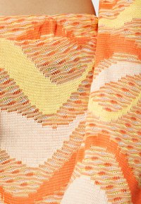 Top em tecido de malha com padrões em zigzag vibrantes em laranja, amarelo e creme, com detalhes texturizados e um design leve e respirável.