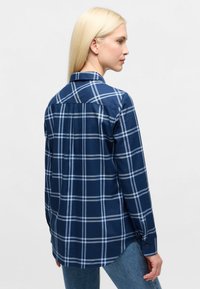 Woman with long blonde hair wearing a blue and white checkered long-sleeve shirt and blue jeans, facing sideways against a plain background.