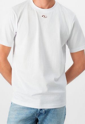 Homme portant un t-shirt blanc uni à manches courtes avec une petite découpe ovale près du col, associé à un jean en denim bleu clair.