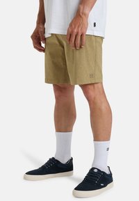 Khaki shorts with a textured pattern, featuring a small logo patch. Paired with navy sneakers and white crew socks.