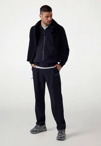 Marineblå atletisk træningsdragt med hættejakke med høj krave og matchende bukser, suppleret med grå sneakers med teksturerede detaljer.