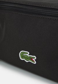 Bolsa de tela negra con un acabado texturizado y un logo de cocodrilo verde con borde blanco y detalles rojos en la parte frontal. Cierre con cremallera.