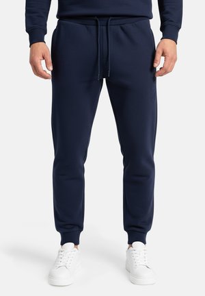 Uomo che indossa pantaloni da jogging blu navy con coulisse in vita e scarpe da ginnastica bianche, in piedi davanti a uno sfondo semplice.