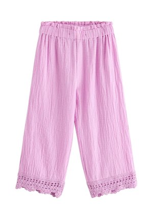 Smilende ung pige med blondt hår, iført en lyserød tekstureret top og bukser med blonderkant og cremefarvede sko, stående mod en ensfarvet baggrund.