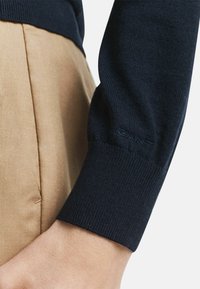 Maglione navy con polsini a costine, abbinato a pantaloni in tessuto beige. Il maglione presenta un marchio discreto sul polsino. Texture liscia.