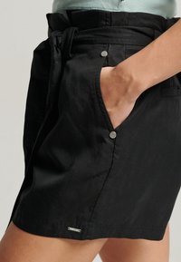 Pantaloni corti neri in tessuto con vita legata, tasche frontali fissate con hardware argentato e un sottile dettaglio sull'orlo. Texture strutturata e liscia.