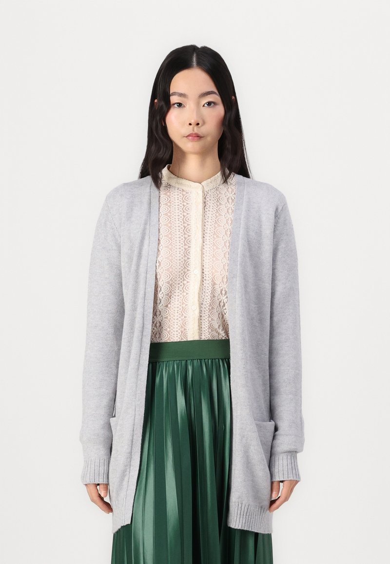 Grå strikket cardigan med åben front, med ribbede manchetter og en lomme. Båret over en mønstret beige bluse og en grøn plisseret nederdel.