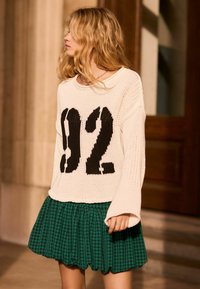 Maglione bianco lavorato a maglia con il numero "92" nero frontale, abbinato a una gonna a quadri verde. Vestibilità ampia, lavorazione testurizzata e orlo svasato.