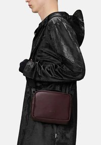Borsa a tracolla in pelle marrone scuro con forma rettangolare, texture liscia, tracolla regolabile e logo in rilievo discreto, indossata contro un cappotto nero.