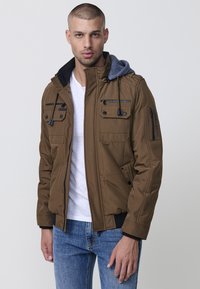 Braune gepolsterte Jacke mit abnehmbarem grauen Kapuzen, Reißverschluss vorne und mehreren Reißverschlusstaschen. Kombiniert mit einem weißen T-Shirt und blauen Jeans.