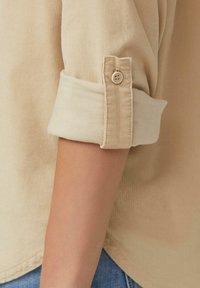 Beige Kordhemd mit aufgerollten Manschetten, das über einen einzelnen Knopfverschluss verfügt. Die Stoffbeschaffenheit wirkt weich und gerippt, mit einem lässigen Schnitt.