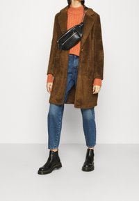 Hnědý, chlupatý kabát přes oranžový žebrovaný svetr, v kombinaci s modrými džíny a černými kotníkovými botami. Černá crossbody taška poskytuje kontrast.