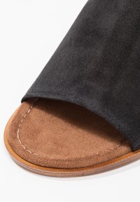 Sandale noire en daim avec une semelle intérieure lisse marron. Elle présente une semelle plate et des coutures visibles le long du bord pour plus de détails.