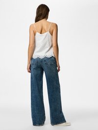 Žena stojící zády k nám, oblečená v bílém krajkovém tílku bez rukávů, modrých širokých džínách a bílých teniskách na jednobarevném pozadí.