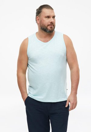 Man met baard en naar achter gebonden haar, gekleed in een lichtblauw mouwloos shirt en donkere broek, staand met handen in de zakken.