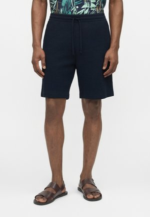 Mann iført sorte teksturerte shorts med snøring og brune lærsandaler, stående foran en ensfarget hvit bakgrunn.