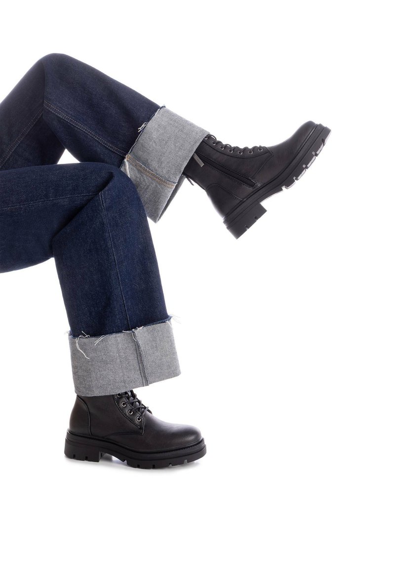 Botas de tobillo negras con cordones, con un upper de cuero texturizado y una suela gruesa, combinadas con jeans de mezclilla oscuros con puños de tela gris.
