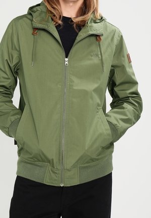 Person trägt eine olivgrüne Kapuzenjacke mit Reißverschluss, die Hände in den vorderen Taschen und schwarze Hose.