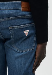 Κοντινό πλάνο του πίσω τσέπα σε μπλε τζιν τζιν με τριγωνικό λογότυπο Guess και καφέ δερμάτινη ετικέτα εμπορικού σήματος πάνω από την τσέπη, χέρι ακουμπισμένο δίπλα.