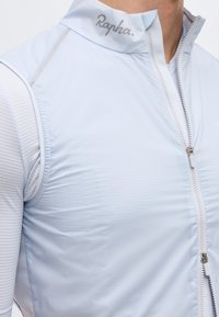 Ανοιχτό μπλε αμάνικο γιλέκο ποδηλασίας με φερμουάρ μπροστά και λογότυπο «Rapha» στον γιακά, φορεμένο πάνω από λευκό ριμπ πουκάμισο με μακριά μανίκια.