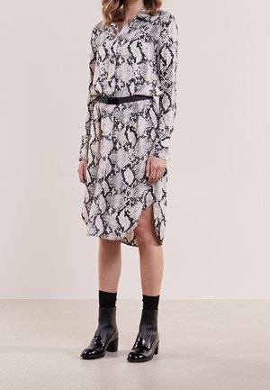Woman wearing a long-sleeve, snake print dress with a black belt and black ankle boots with socks standing against a plain background.