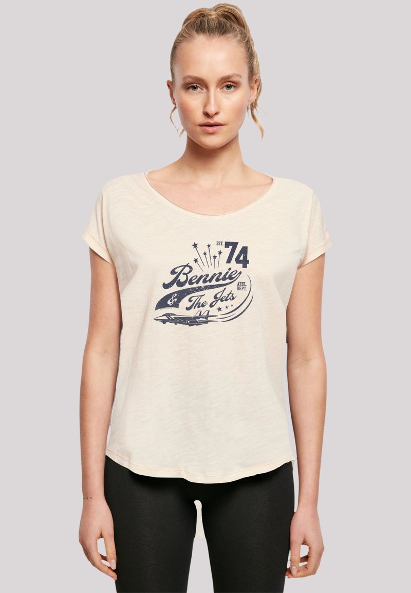Model trägt ein hellcremfarbenes T-Shirt mit einem dunkelblauen grafischen Design, das "Bennie & The Jets" und ein Flugzeug zeigt, kombiniert mit schwarzen Leggings.