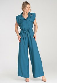Turkusowy kombinezon z kołnierzykiem, frontowymi guzikami i krótkimi, usztywnionymi rękawami. Posiada pasek w talii oraz szeroką nogawkę. Gładka tekstura materiału.