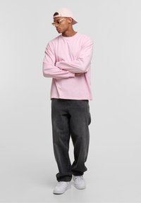 Lichtroze longsleeve shirt, grijze baggy jeans, witte sneakers en een lichtroze cap; heeft duidelijke lijnen en een casual pasvorm.