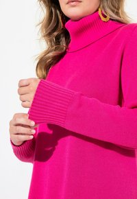 Klarr rosa polotröja med ribbad krage och manschetter; slät textur. Modellens händer greppar ärmen, vilket visar passform och design detaljer.