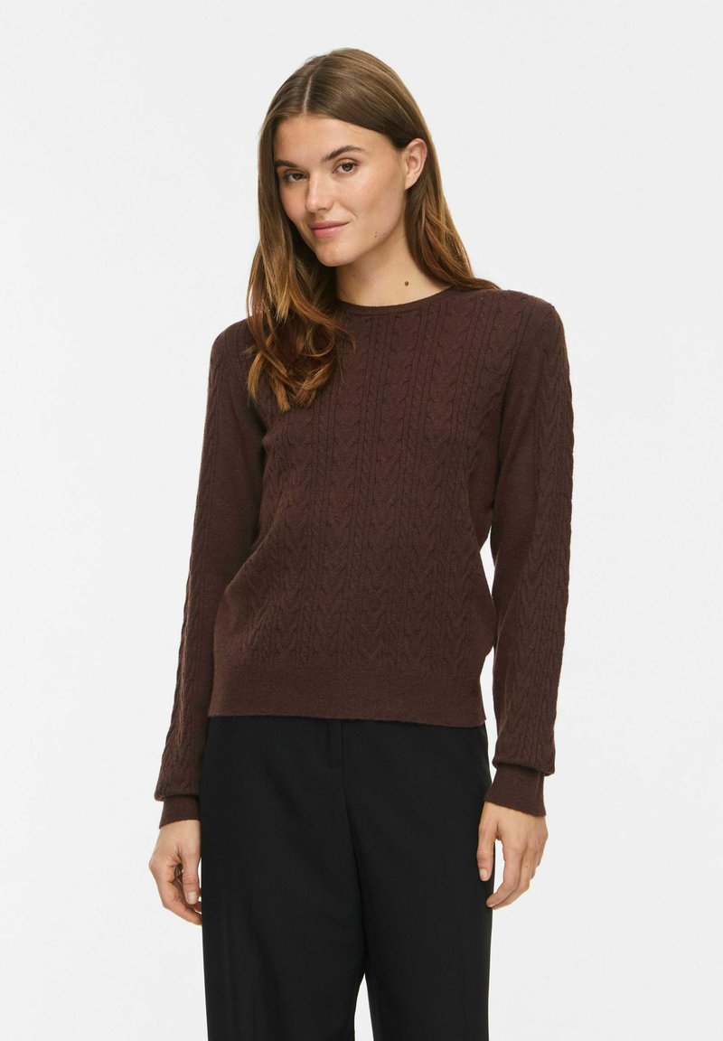 Femme aux longs cheveux bruns portant un pull torsadé marron foncé et un pantalon noir, debout devant un fond clair uni.