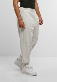 Muž v svetlosivých teplákoch, bielych ponožkách a bielych teniskách Nike stojí pred jednoduchým sivým pozadím.