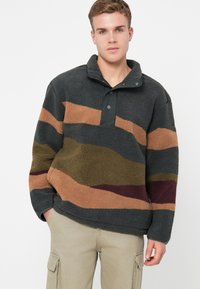 Fleece-Pullover in Dunkelgrau mit hellbraunen, olivgrünen und burgunderroten wellenförmigen Streifen; verfügt über eine schwarze Druckknopfleiste und strukturierter Stoff.