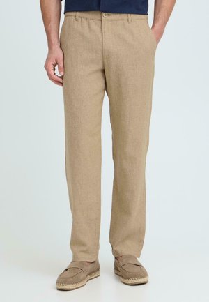 Hombre con pantalones de lino beige con bolsillos laterales y esparteñas beige a juego, de pie contra un fondo claro y liso.