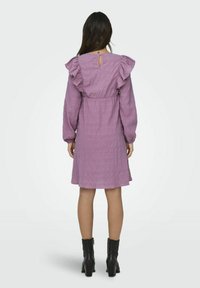 Robe violette à volants avec des manches longues, en tissu texturé, et un dos en keyhole. Portée avec des bottines noires à la cheville.