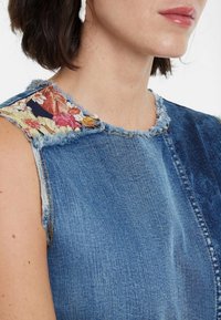 Top in denim con orli sfrangiati e tessuto a motivi floreali sulle spalle. Presenta una base blu chiaro con accenti scuri.