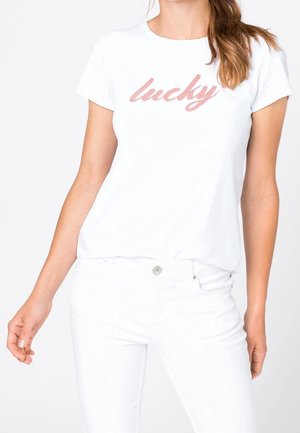 Frau trägt weißes T-Shirt mit kurzen Ärmeln und pinkem "lucky"-Schriftzug sowie weiße Hose, stehend vor einem schlichten weißen Hintergrund.