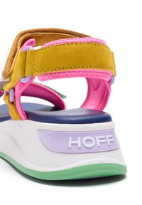 Mångfärgad sandal med ett gult mockaband, rosa detaljer, en lila logotyp och en kraftig vit sula med gröna inslag.