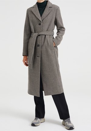 Manteau long à motifs en beige et noir, doté d'un col cranté, de boutons double boutonnage, d'une ceinture et de poches latérales, porté sur un pantalon sombre.
