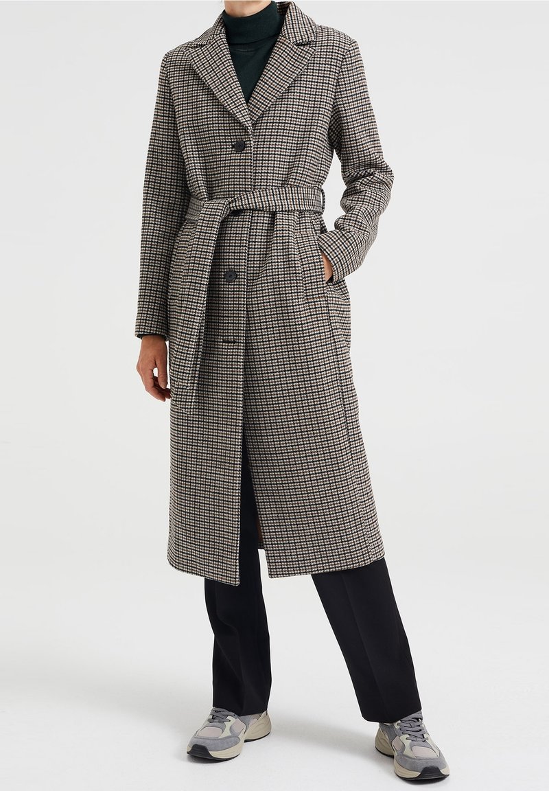 Lång, mönstrad kappa i beige och svart, med skört krage, dubbelknäppning, bälte och sidofickor, bärs över mörka byxor.