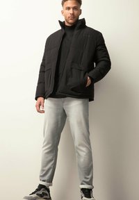 Sort polstret jakke med høj krave, der har to frontlommer. Båret over en sort skjorte, parret med lysegrå jeans og sorte sneakers.