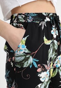 Pantalon à motifs floraux noirs avec des feuilles vertes et des fleurs bleues, doté d'une ceinture élastique et de poches latérales. Texture de tissu douce.