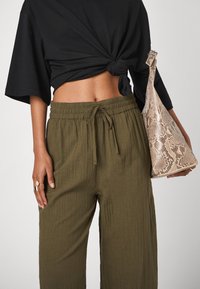 Haut à manches courtes noir avec un nœud, pantalon large plissé vert olive avec une ceinture élastique, et un sac à main léger avec imprimé serpent.