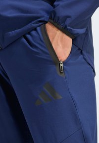 Granatowe spodnie sportowe z czarną kieszenią na zamek błyskawiczny, elastycznym pasem i widocznym czarnym logo Adidas po boku. Gładki, lekki materiał.