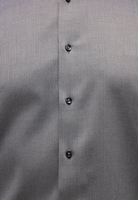 Button-up Hemd mit schwarz-weißer geometrischer Musterung, präsentiert sich mit einer glatten Stoffoberfläche und sieben schwarzen Knöpfen auf der Vorderseite.