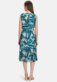 Bezrękawna sukienka w odcieniach teal i beżowym, z plisami oraz liściastym wzorem, wcięcie w talii, zwiewny dół i subtelna textura. Widok z tyłu ukazuje otwarty dekolt.