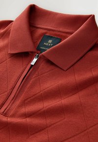 Polo rouge à manches courtes avec un motif en diamant texturé, fermeture éclair et col côtelé. Présente une étiquette noire avec "NEXT" en doré.