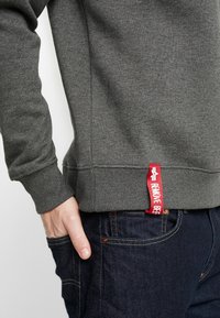 Sudadera gris oscuro hecha de un material texturizado suave, con una etiqueta roja que dice "REMOVE BEF" en el dobladillo inferior.