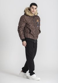 Giubbotto imbottito marrone con cappuccio foderato in pelliccia, caratterizzato da un motivo a logo ripetuto e un patch a cartone animato. Indossato con pantaloni neri e sneaker bianche.