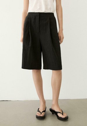 Femme portant un short large noir taille haute à plis, arrivant au genou, un haut blanc et des sandales noires à talons, debout sur un sol gris.